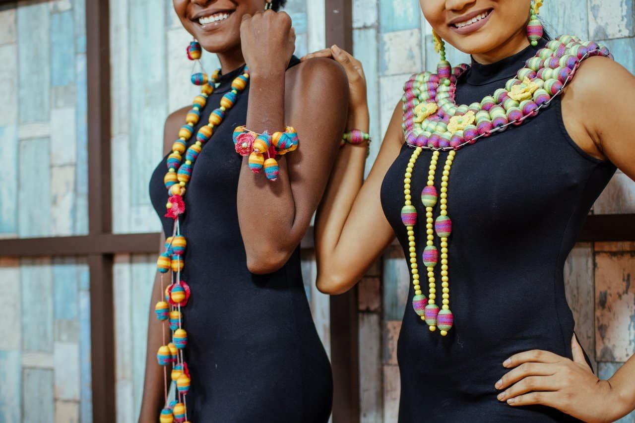 Two African models showcasing vibrant beaded jewelry and elegant fashion against a rustic backdrop.